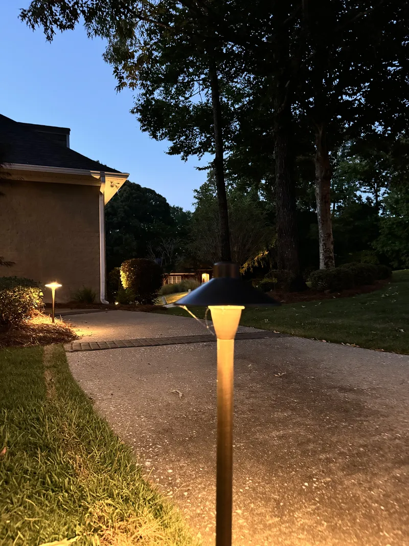Brass path light glowing beside a driveway with a lit home behind at dusk